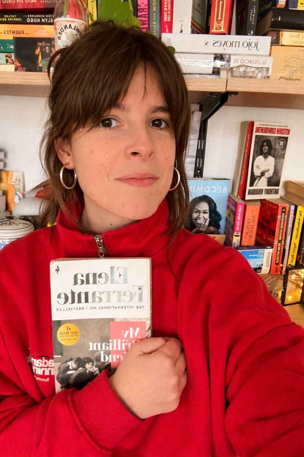 Paule is standing in front of a bookshelf, holding "My brilliant friend" by Elena Ferrante.